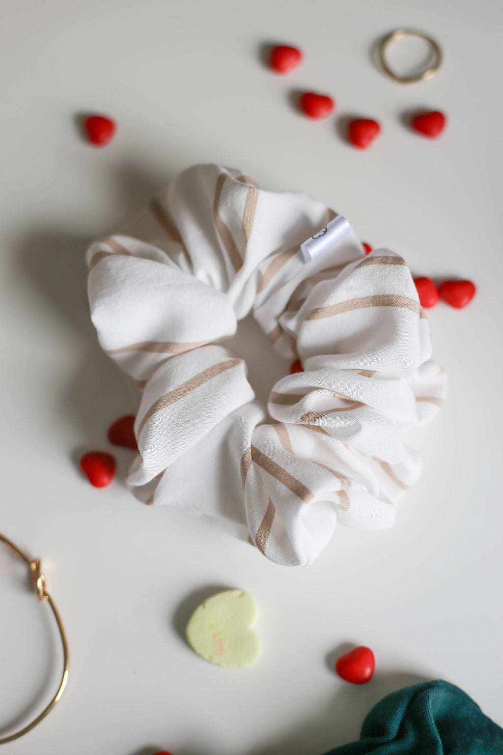 White scrunchie with gold stripes on a light gray background with small red hearts and gold rings.
