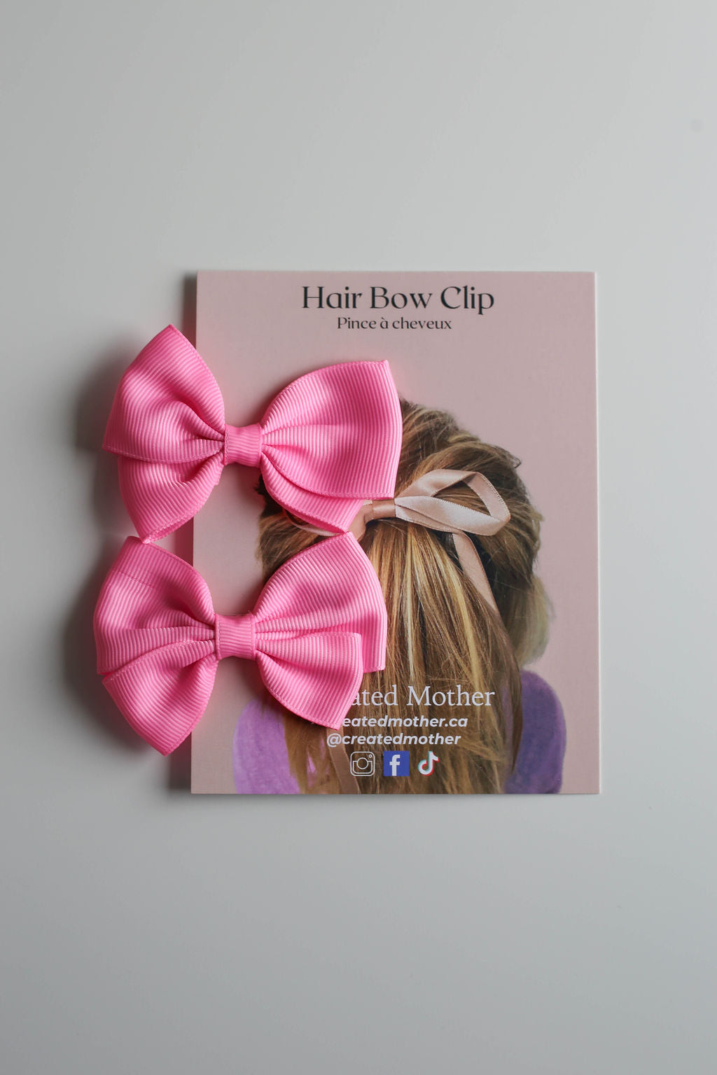 Small ribbon bow hair clip in pastel pink, attached to a metal alligator clip and displayed on a branded card.