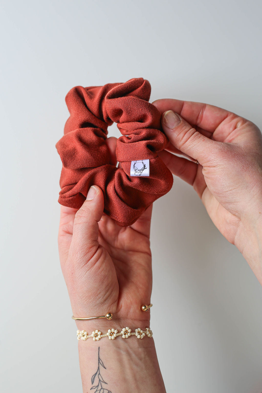 Rust scrunchie held by a hand with gold jewelry on a light gray background