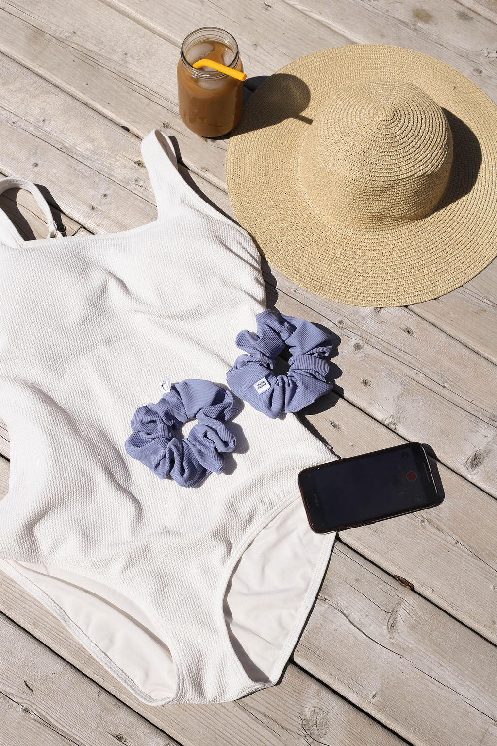 White swimsuit, straw hat, purple scrunchies, and smartphone on a wooden surface