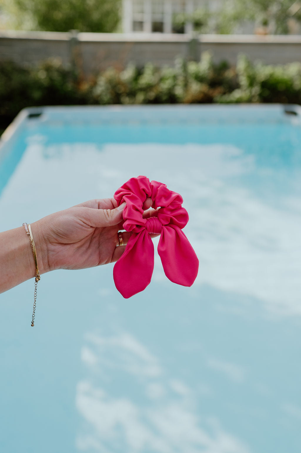 Hand holding a pink SWIM scrunchie with a pool and garden in the background