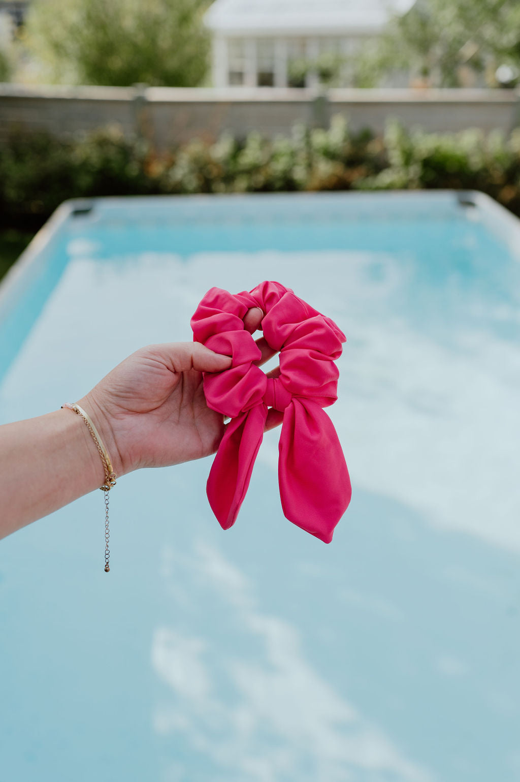 Hand holding a large pink scrunchie with a removable bow, a pool and garden are in the background