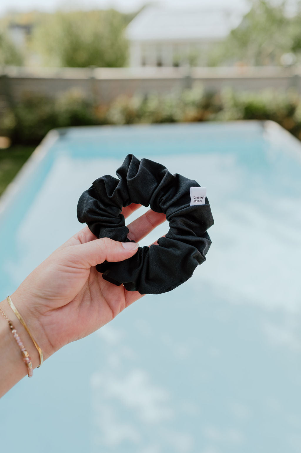 Black scrunchie held by a hand with a blurred pool and greenery in the background
