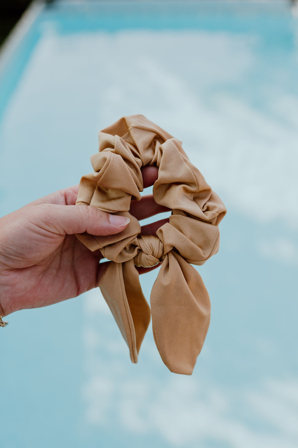 Brown scrunchie with a bow held by a hand against a blurred pool background