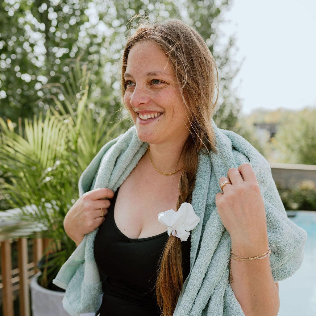 Woman in a black swimsuit with a green towel draped over her shoulders, standing outdoors with a SWIM Scrunchie