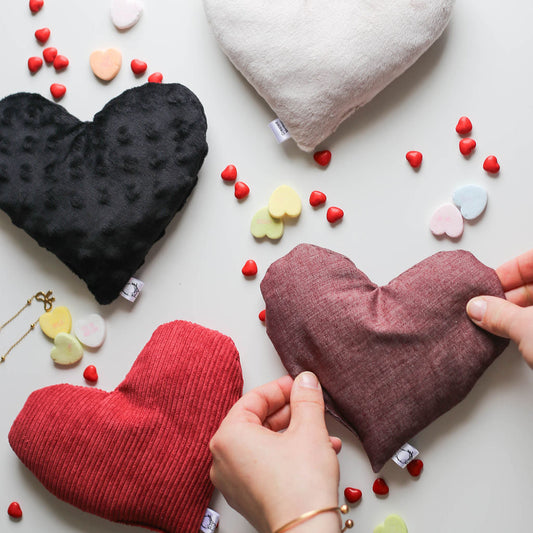Heart-shaped hot cold therapy packs in black, red, and pink on a light surface with small colorful objects.