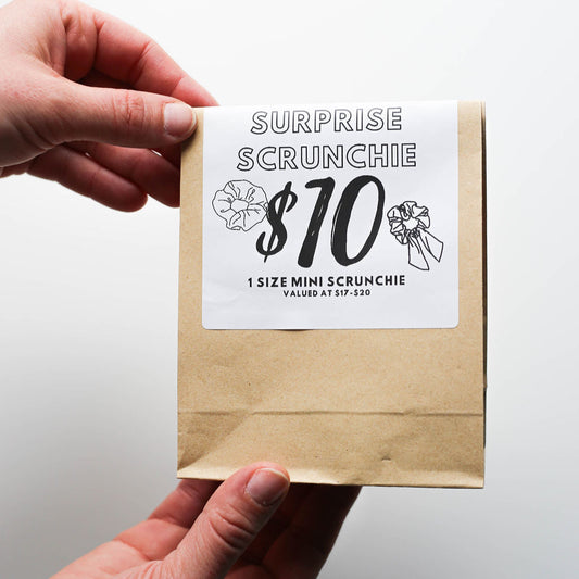 Hand holding a brown paper bag with a surprise scrunchie label on a white background