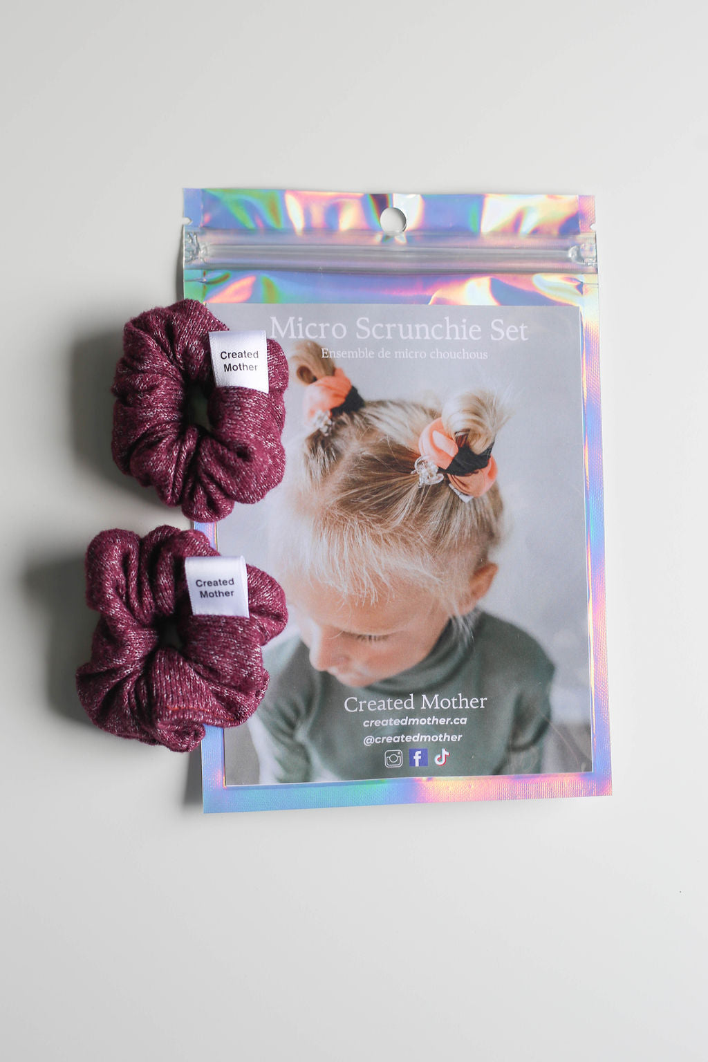 Two maroon scrunchies with packaging featuring a child's face and 'Created Mother' branding on a gray background.
