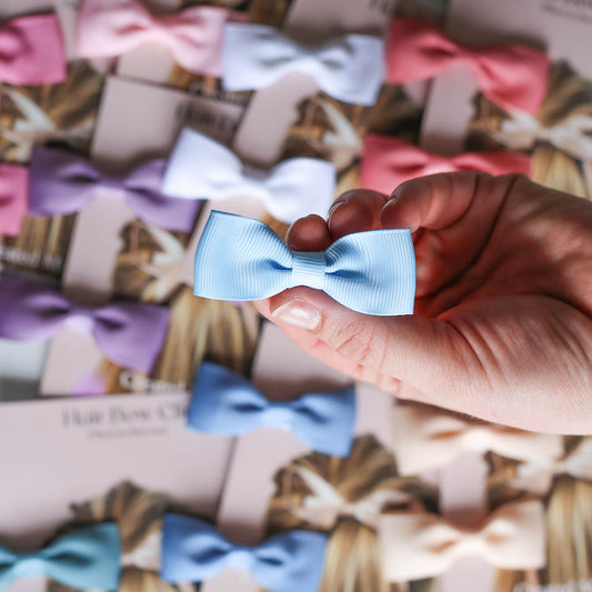 Flat lay of assorted small bow clips in pastel tones, each set on branded packaging.