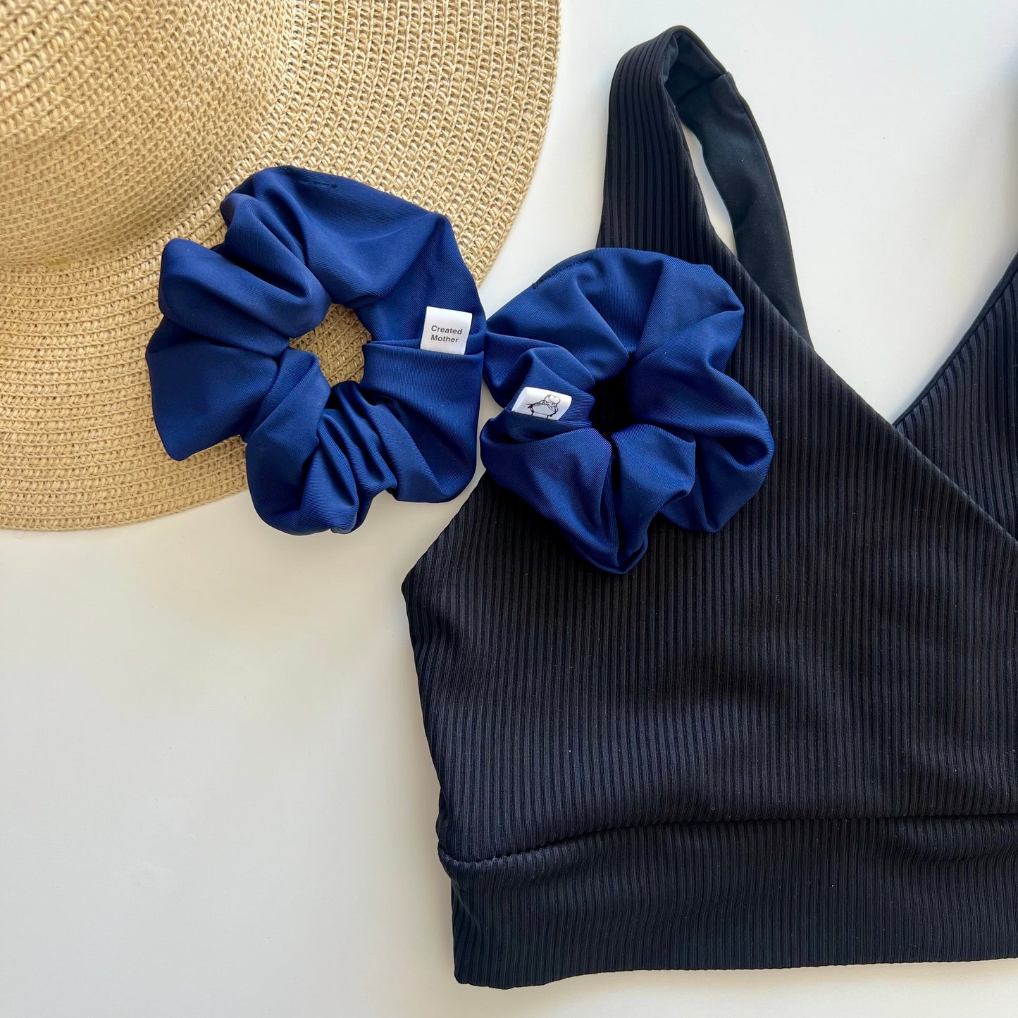 Navy blue scrunchies and black bikini top on a white surface with a straw hat.