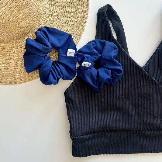 Navy blue scrunchies and black bikini top on a white surface with a straw hat.