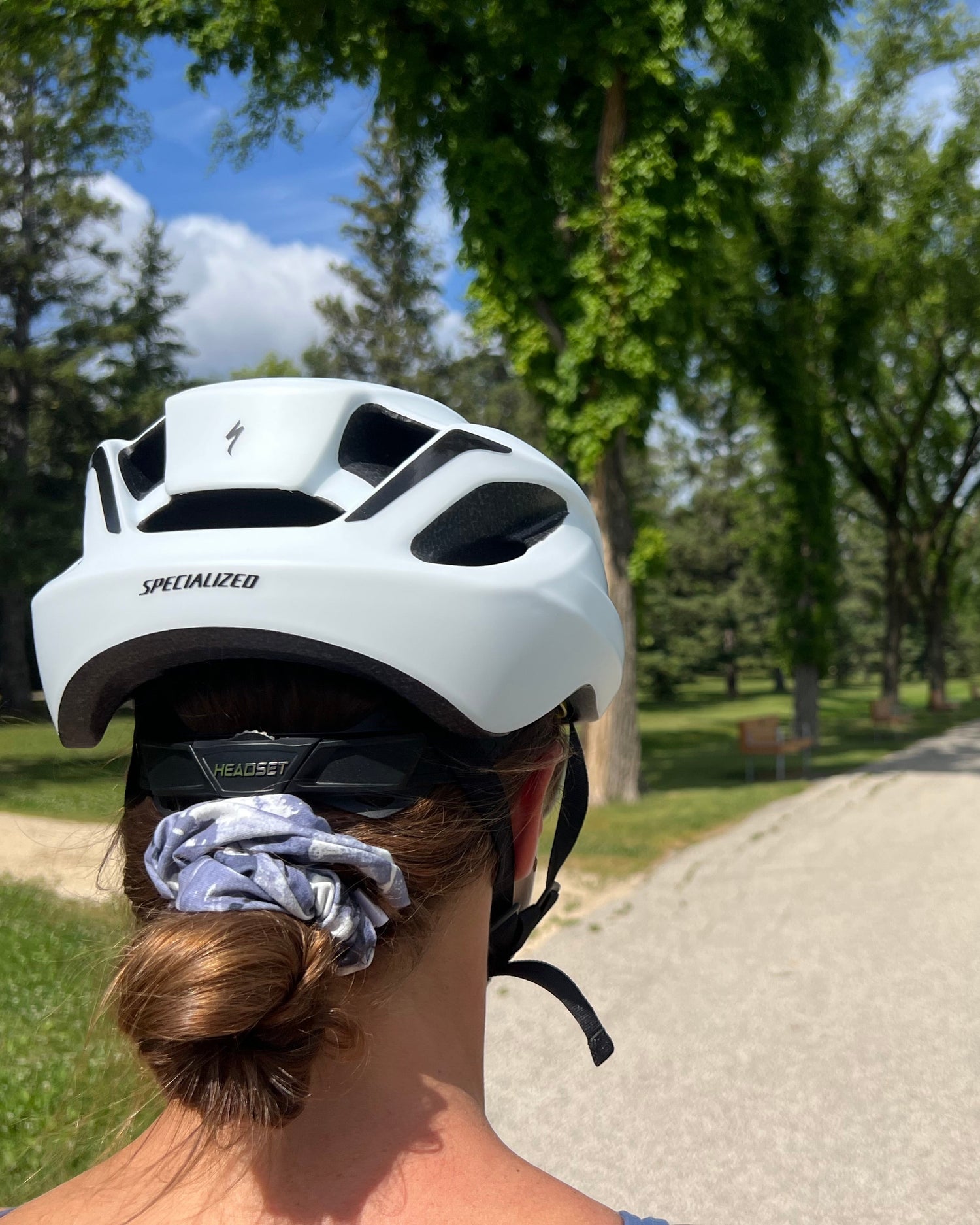 Watercolour sport scrunchie with a white Specialized cycling helmet worn by a person seen from behind, riding on a paved path in a park with trees and blue sky.