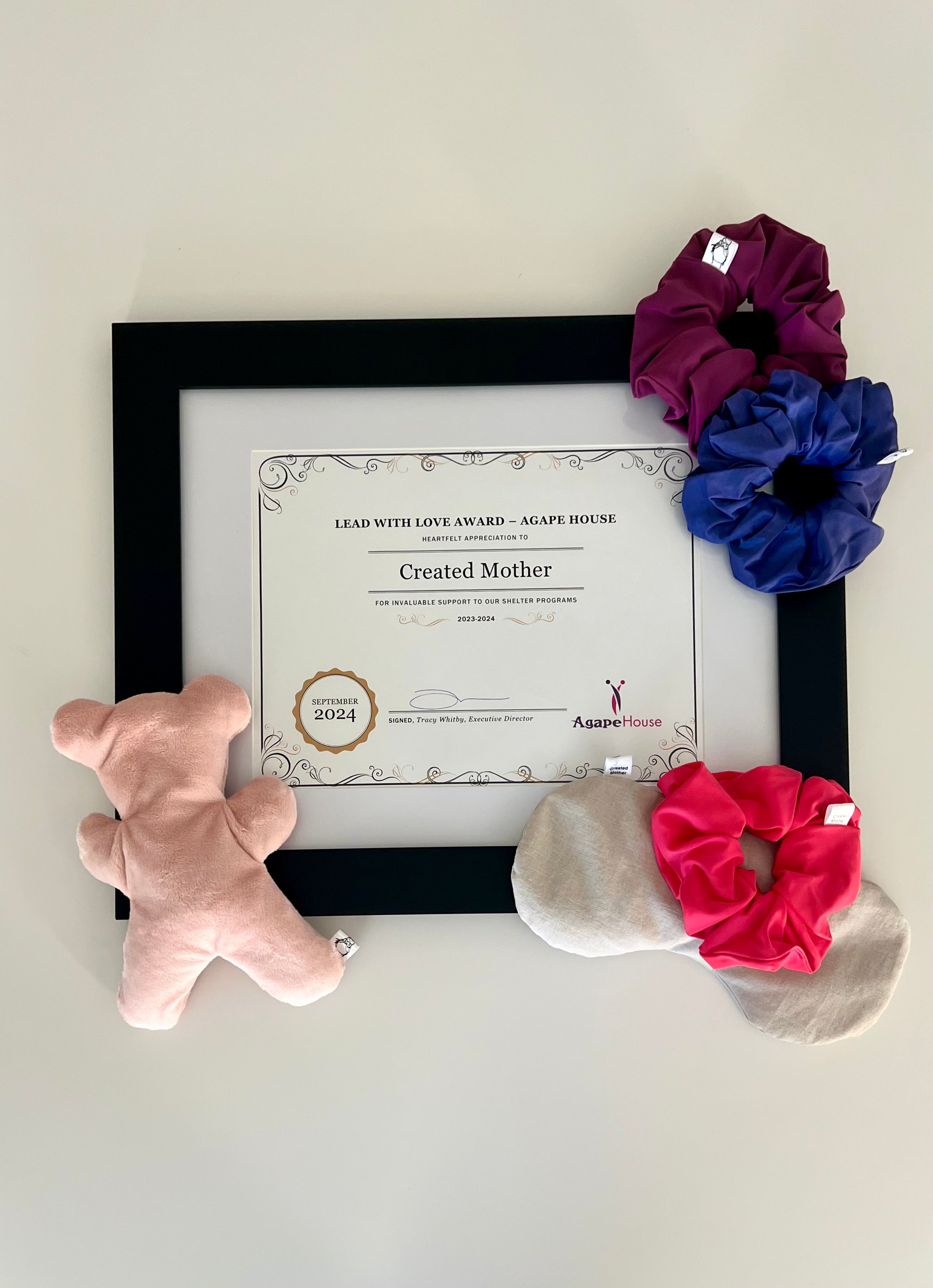 Framed certificate with created mother products including a therapy bear bear, scrunchies, and an eye mask on a light background.