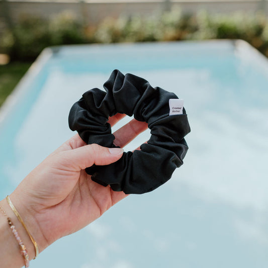 Black scrunchie held by a hand with a blurred pool and greenery in the background