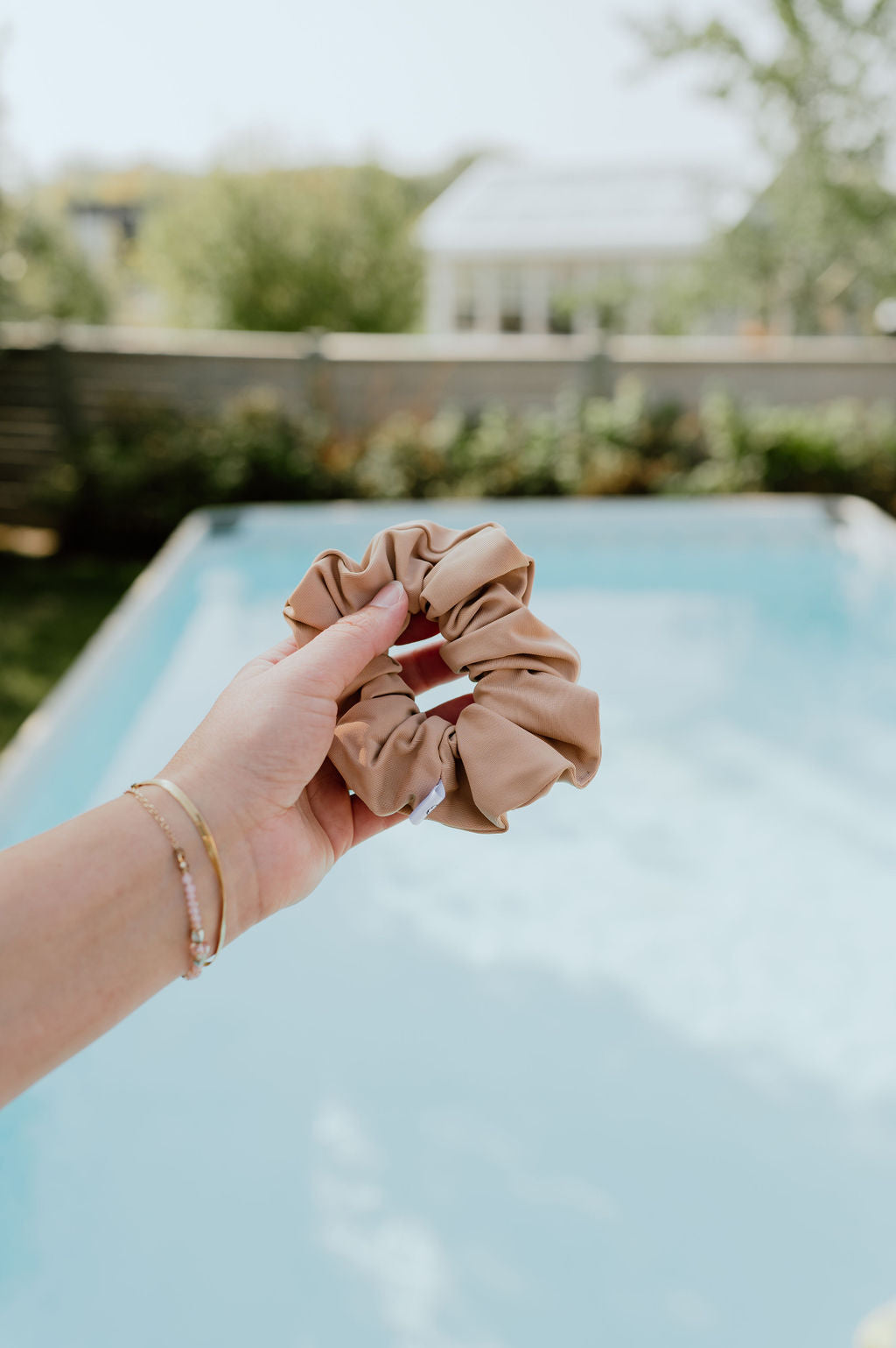 Hand holding a beige scrunchie with a pool and garden in the background