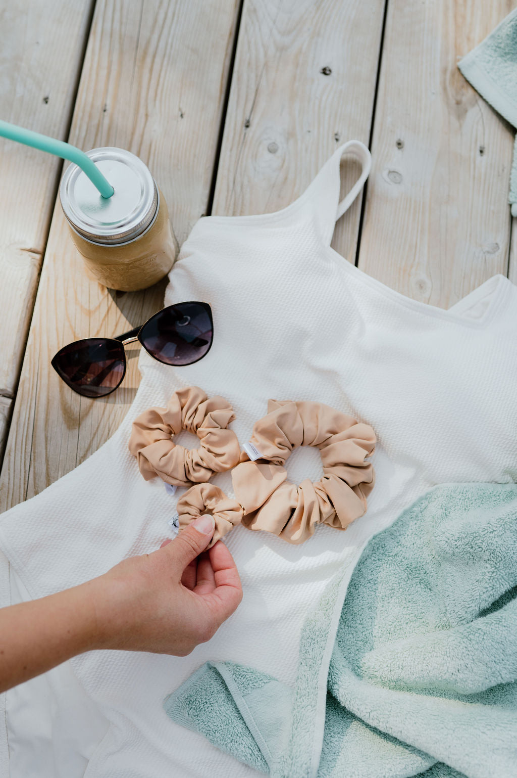 Three beige scrunchies on a white towel with sunglasses and a drink on a wooden surface.