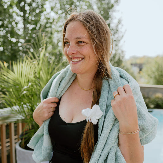 Woman in a black swimsuit with a green towel draped over her shoulders, standing outdoors with a SWIM Scrunchie